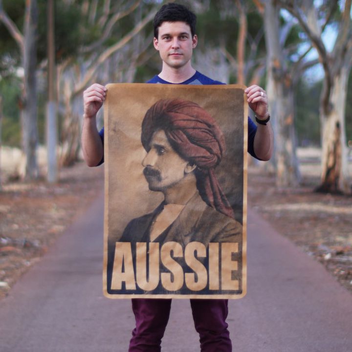 Photograph of a man standing holding a large poster. The poster depicts the portiat of a man wearing a red turban with a moustache and the word aussie appears in large text across the bottom